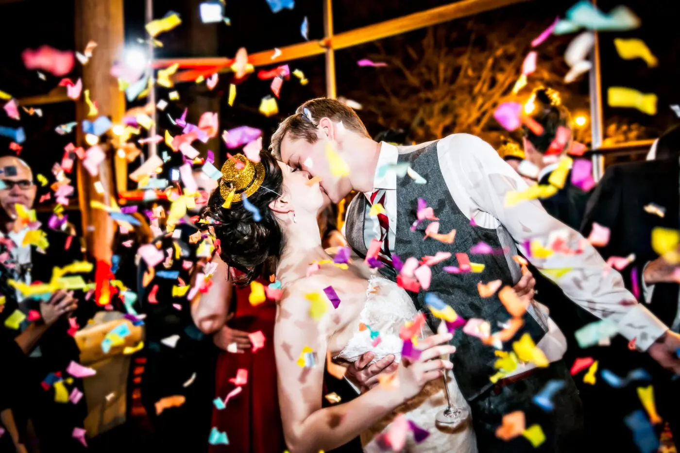 A bride and groom kiss as colorful confetti falls through the air around them and their surrounding guests.