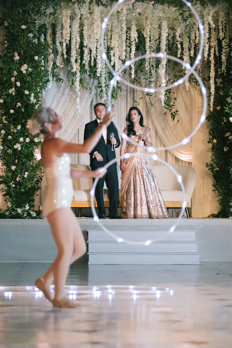 A person in a dazzling outfit performs a captivating hoop routine with three hoops in front of a couple. The couple, dressed in formal attire, stands on a white platform under an arch of hanging flowers and greenery, watching the performance.