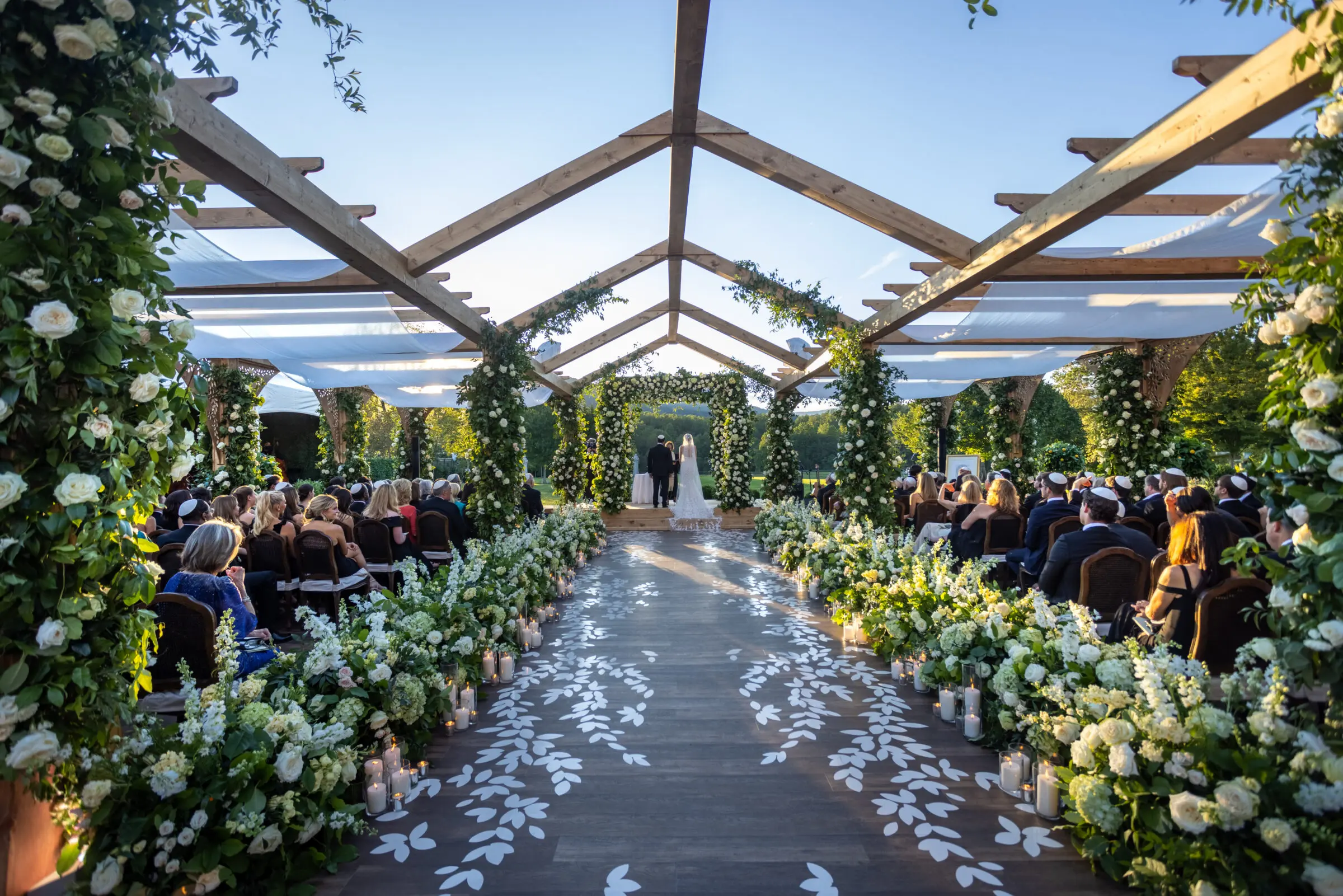 A beautifully decorated outdoor wedding aisle with a wooden pergola draped and green foliage filled with white flowers shows a bride and groom stand at the altar. The aisle is lined with white floral arrangements and illuminated with candles, leading to a flower-adorned altar.