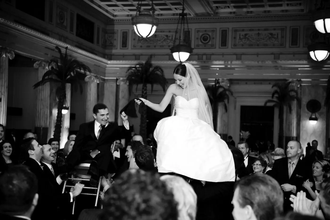 A bride and groom are lifted on chairs by guests in a grand, ornately decorated venue during their wedding celebration. The bride, in a strapless gown and veil, and the groom, in a suit, hold hands and smile joyfully. Friends and family surround them, clapping and cheering.