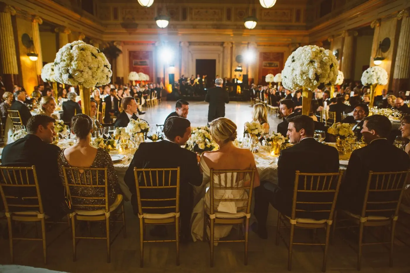 A formal event in a grand hall with chandeliers. Guests in elegant attire sit at tables adorned with white floral centerpieces. The focus is on a group seated at a long table with their backs to the camera. A band plays in the background.