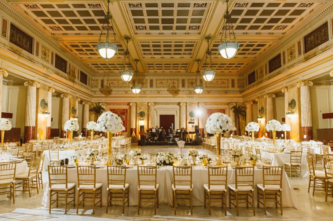 A grand hall is decorated with long tables in white tablecloths, gold chairs, and tall white florals. A band is set up in the background on a stage.