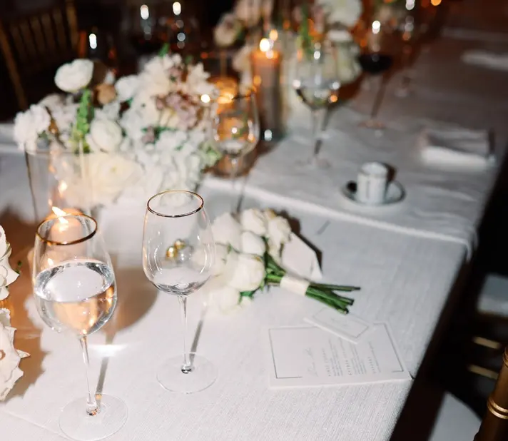 A wedding reception table with white tablecloth, adorned with elegant floral arrangements, wine glasses, and lit candles. The romantic ambiance is enhanced by the warm glow of lights in the background. A wedding program and table setting are also visible.
