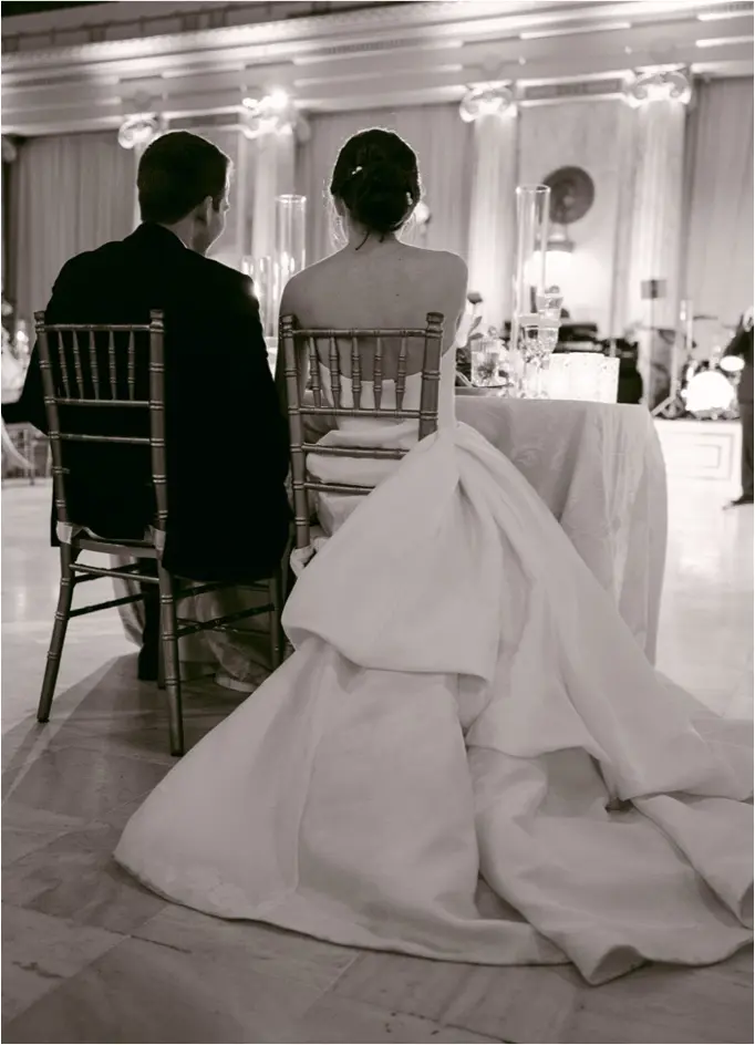 A bride and groom sit at the wedding reception, backs turned to the camera. The bride wears a long strapless gown and the groom wears a dark suit. The moment is captured in black and white.