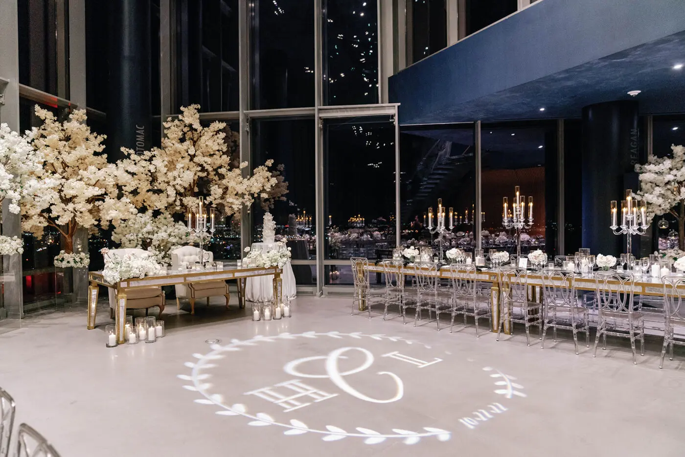 A modern wedding reception venue with floor-to-ceiling windows showcasing a nighttime view. White florals and candles adorn long banquet tables. A personalized monogram is projected on the polished floor, surrounded by a delicate wreath design.