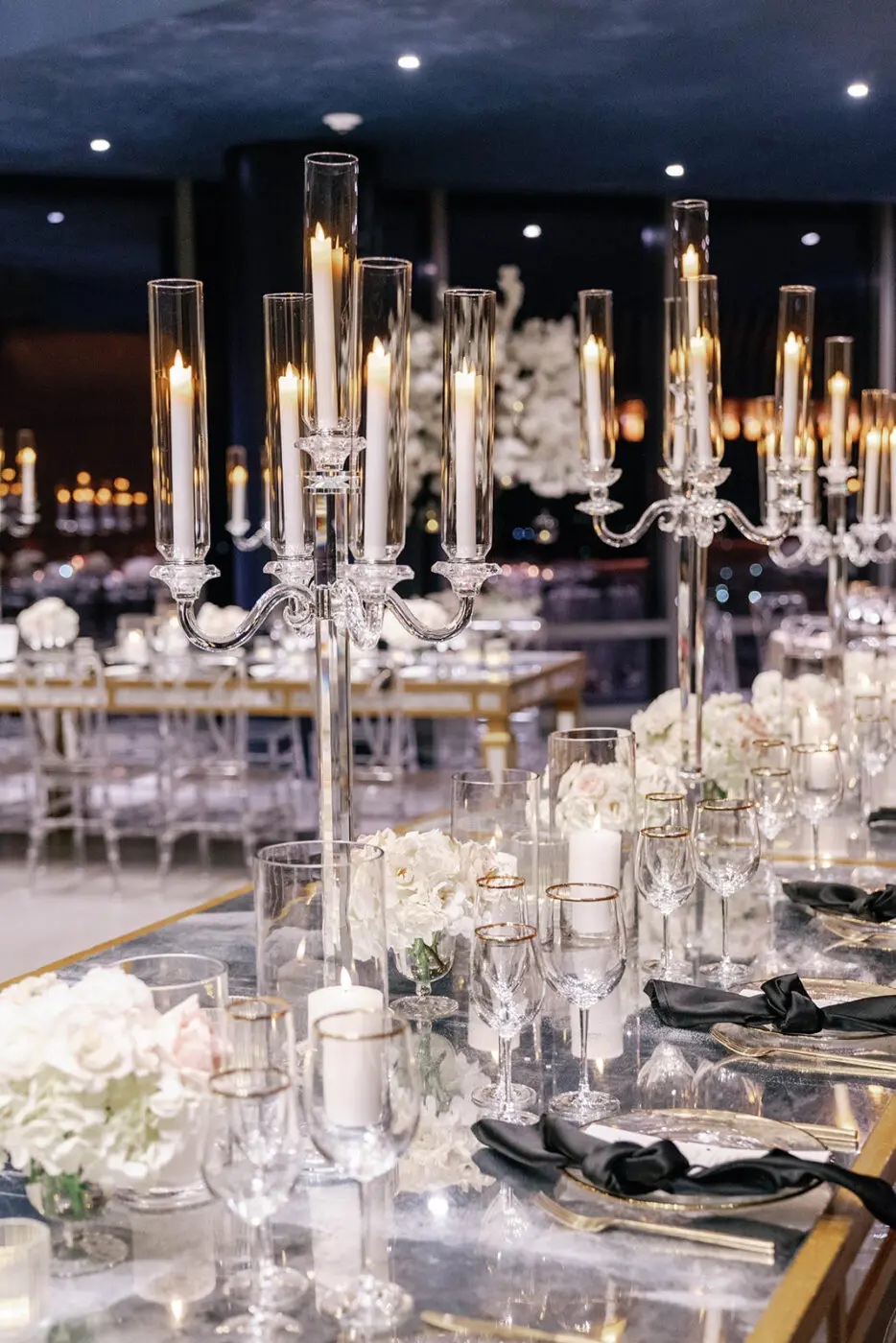 Glass candelabras sit on a decorated table adorned with white florals and glassware.