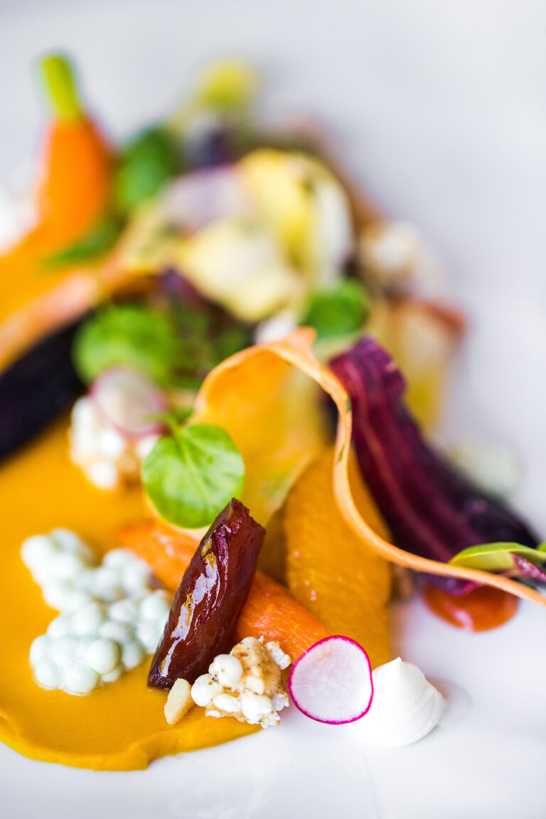 A colorful gourmet vegetable dish is artfully arranged on a plate. The dish includes slices of carrots, radishes, leafy greens, and other vegetables, garnished with dollops of creamy white sauce and small, crispy grains, set against a vibrant orange puree.