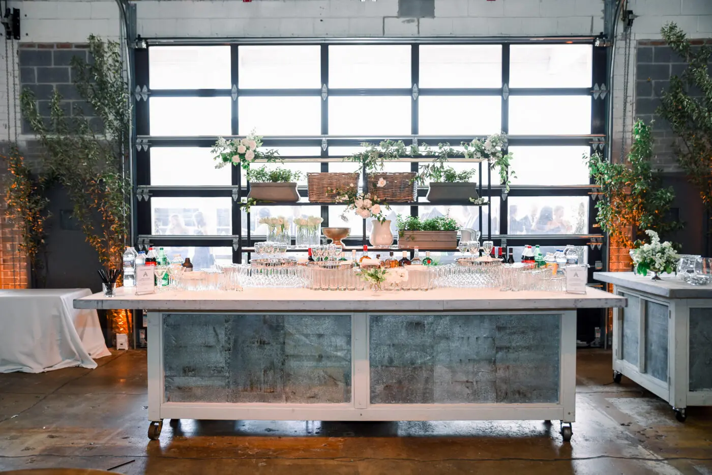 A drink station at a reception features bottled drinks, glassware, white florals, and greenery on an industrial-style white-and-gray buffet table.