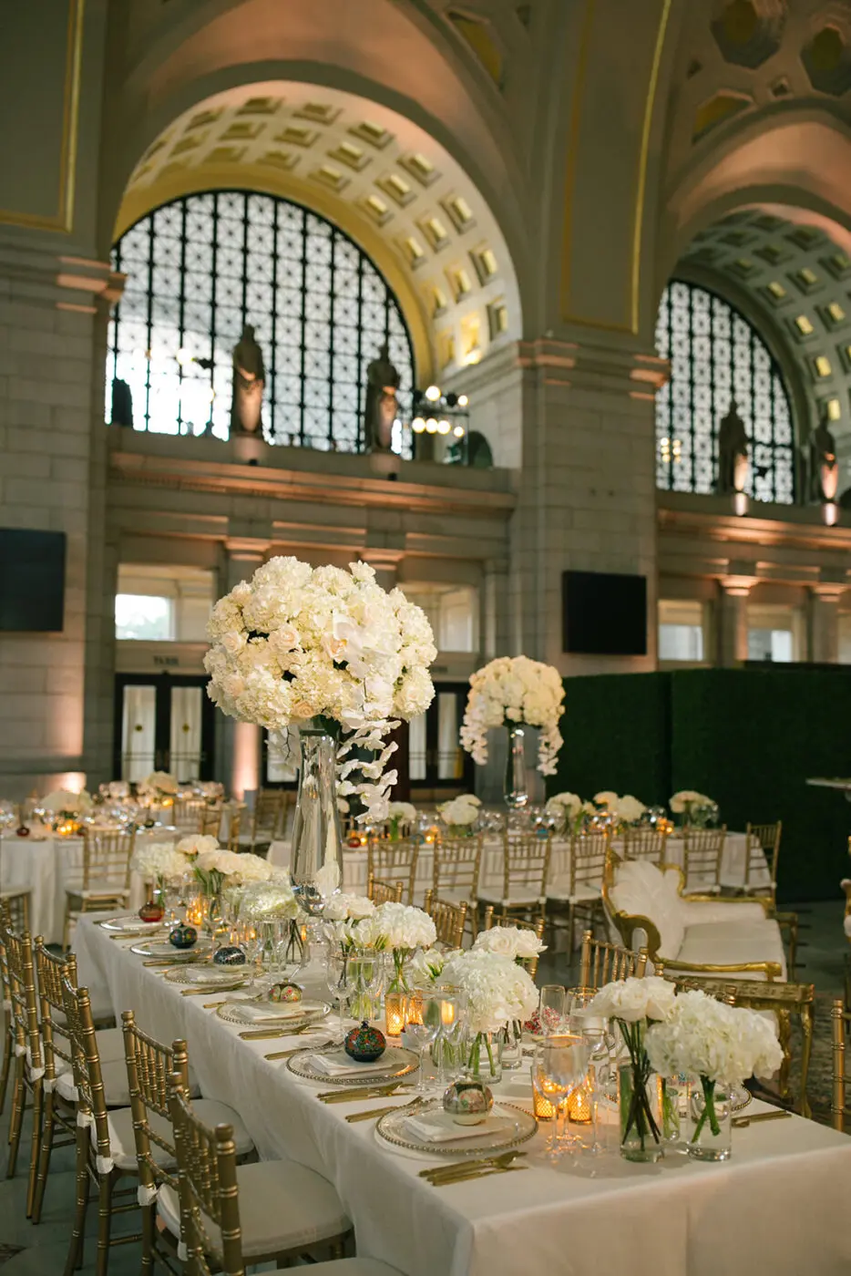 An elegant banquet hall featuring long tables adorned with white floral centerpieces and gold accents. Tall arrangements of white flowers in crystal vases stand on the tables. The hall has high arched windows, intricate decor, and warm ambient lighting.