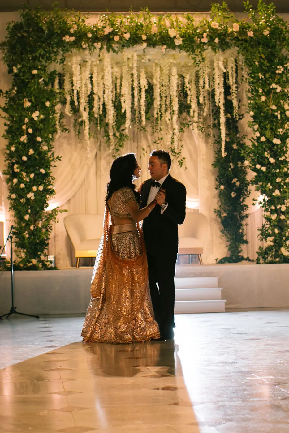 A couple shares a dance in an elegant setting. The woman is wearing a gold legenga and the man is dressed in a black tuxedo. They are surrounded by lush green and white floral arrangements hanging from above, with soft lighting casting a warm glow.