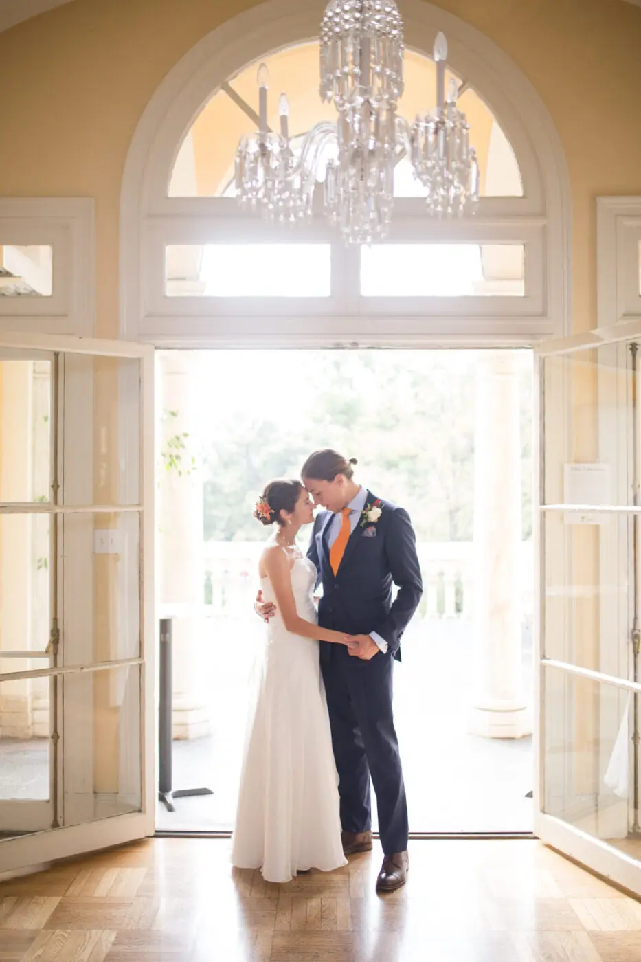 A bride and groom are standing close together in front of a large, arched window with glass doors. The bride is wearing a white gown with flowers in her hair, while the groom is in a dark suit with an orange tie.