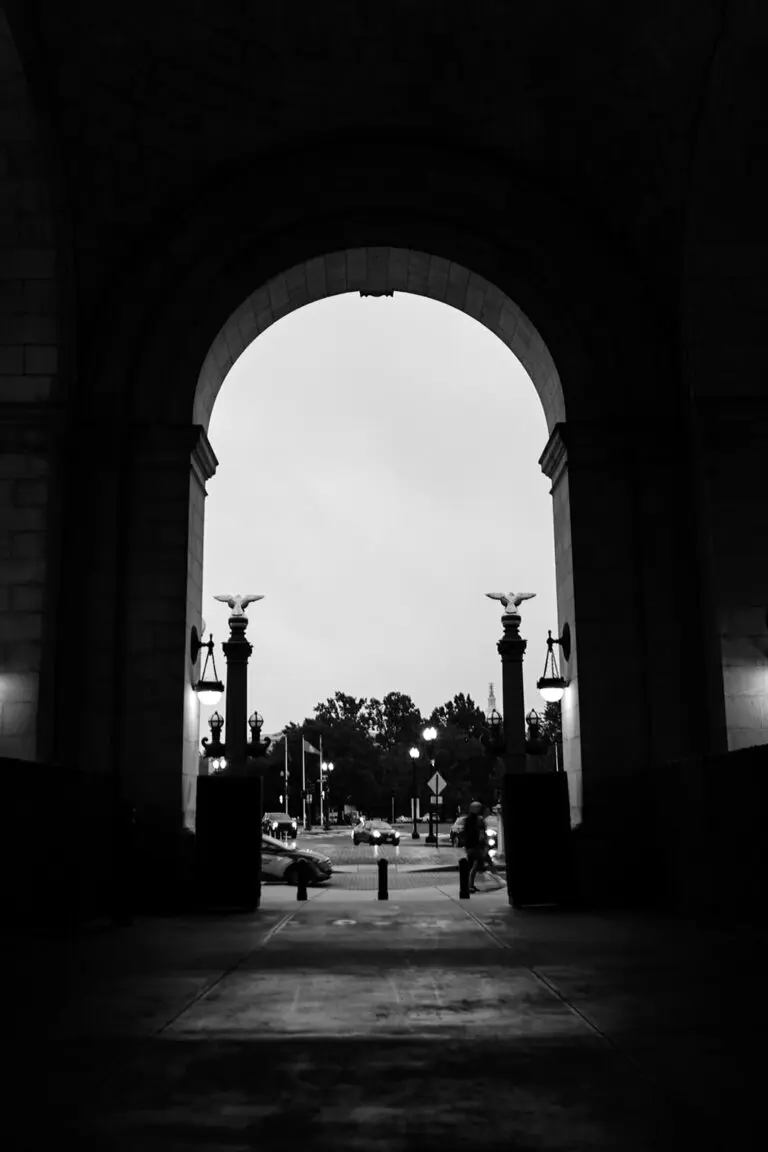 Black and white photo of a large archway with pedestrians and cars visible outside. The arch is adorned with two lantern-style lights and decorative elements. Some streetlights and trees are visible in the background, contributing to the urban setting.