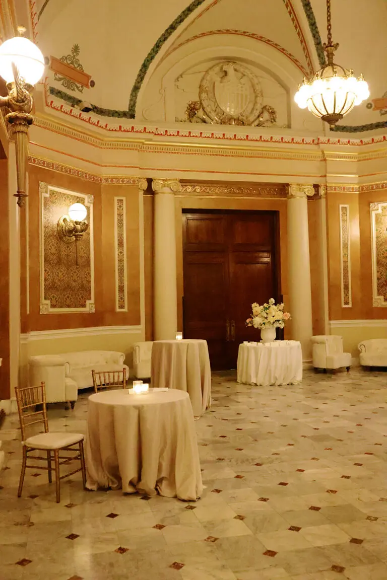 A grand, elegant room with high ceilings and intricate moldings features round tables covered with beige tablecloths, chairs, and floral centerpieces. Sconces and chandeliers illuminate the space, and large wooden doors are seen in the background.