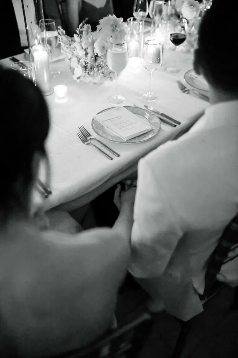 A black-and-white image of a couple sitting at a beautifully set dining table, holding hands. The table is adorned with wine glasses, candles, and floral arrangements, creating an elegant and intimate atmosphere. The couple’s backs are facing the camera.