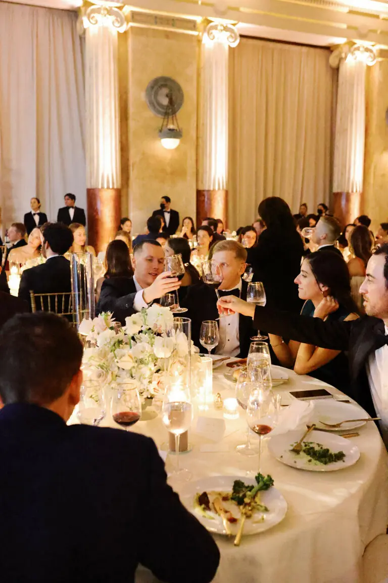 A group of people in formal attire are sitting around a table at an event. They are raising wine glasses in a toast, surrounded by candlelight and floral centerpieces. The room is decorated with large columns and soft lighting, and other guests are in the background.