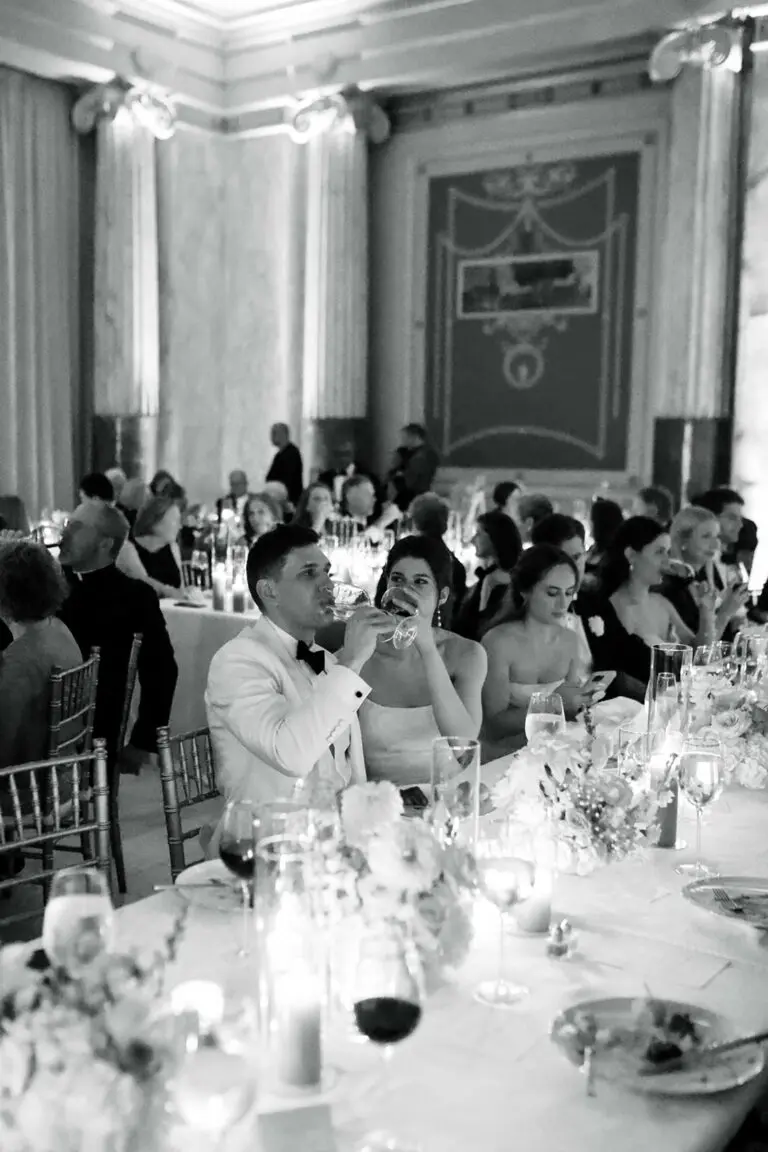 A black and white photo of an elegantly decorated banquet hall filled with guests seated at long tables adorned with floral arrangements and candles. In the foreground, a couple in formal attire is seated and drinking from wine glasses.