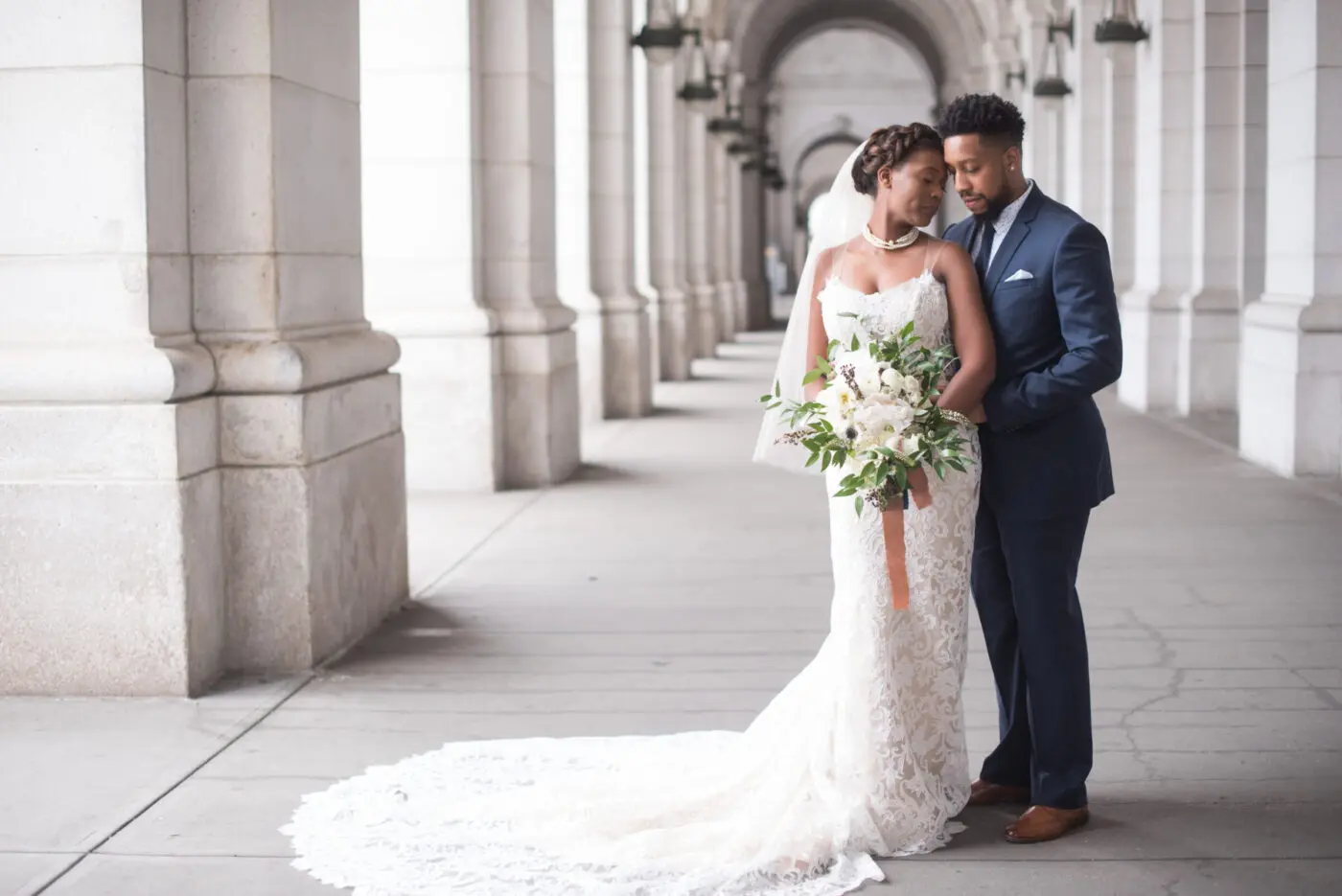A bride and groom pose under an archway outside Union Station. The bride wears a veil and holds a bouquet of white flowers and greenery. The bride wears a white dress and the groom wears a dark blue suit.
