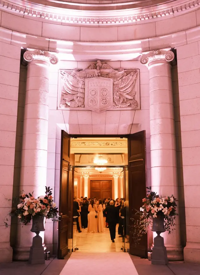 A grand entrance with tall wooden doors opens to an ornately decorated interior. Flower arrangements flanking the door are illuminated by soft, pink lighting. Inside, a chandelier hangs above a large group of people, dressed in formal attire.