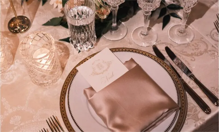 Elegant table setting featuring crystal glassware, silver cutlery, and a white plate with a gold rim. Surrounding the plate are flower arrangements and lighted candles, set on a white patterned tablecloth.