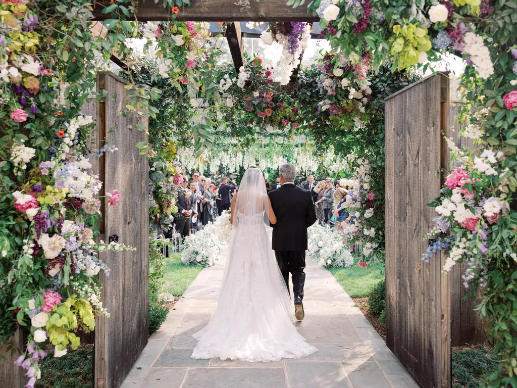 A bride in a white gown and veil walks down an aisle with an older man through an archway adorned with vibrant flowers. They approach a crowd of seated guests, with greenery and floral decorations hanging above and surrounding them. The setting is outdoors.