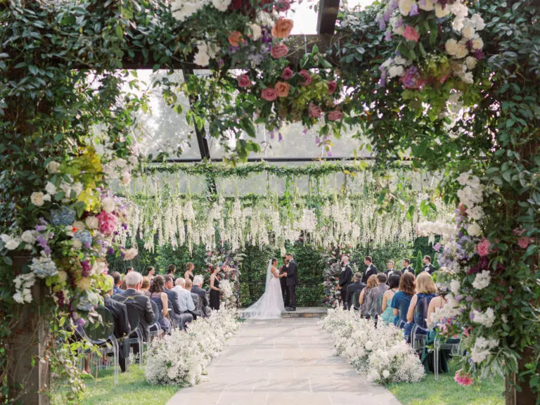 An outdoor wedding ceremony with a lush floral arch framing the couple as they exchange vows. The aisle is lined with white flower arrangements, and guests are seated on either side, surrounded by greenery and colorful hanging floral decorations.