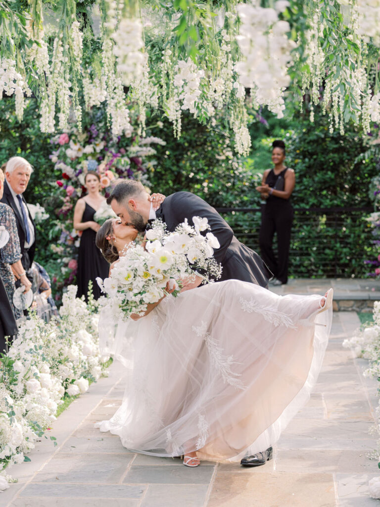 A newlywed couple shares a kiss beneath hanging white flowers and lush greenery. The bride's gown flows as the groom dips her. Flowers line the aisle, and an outdoor lush garden setting enhances the atmosphere.