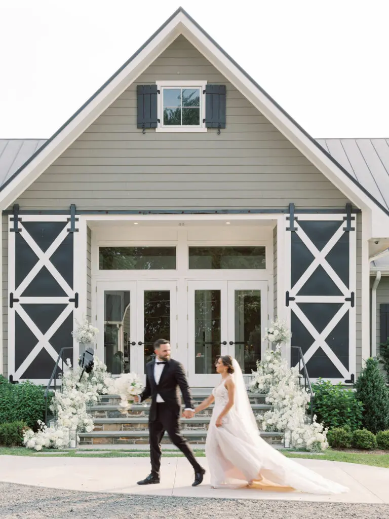 A couple walks hand-in-hand in front of a modern barn-style building. The bride wears a flowing white gown and veil, while the groom wears a black tuxedo. White floral arrangements adorn the steps leading to a set of double doors with large windows.