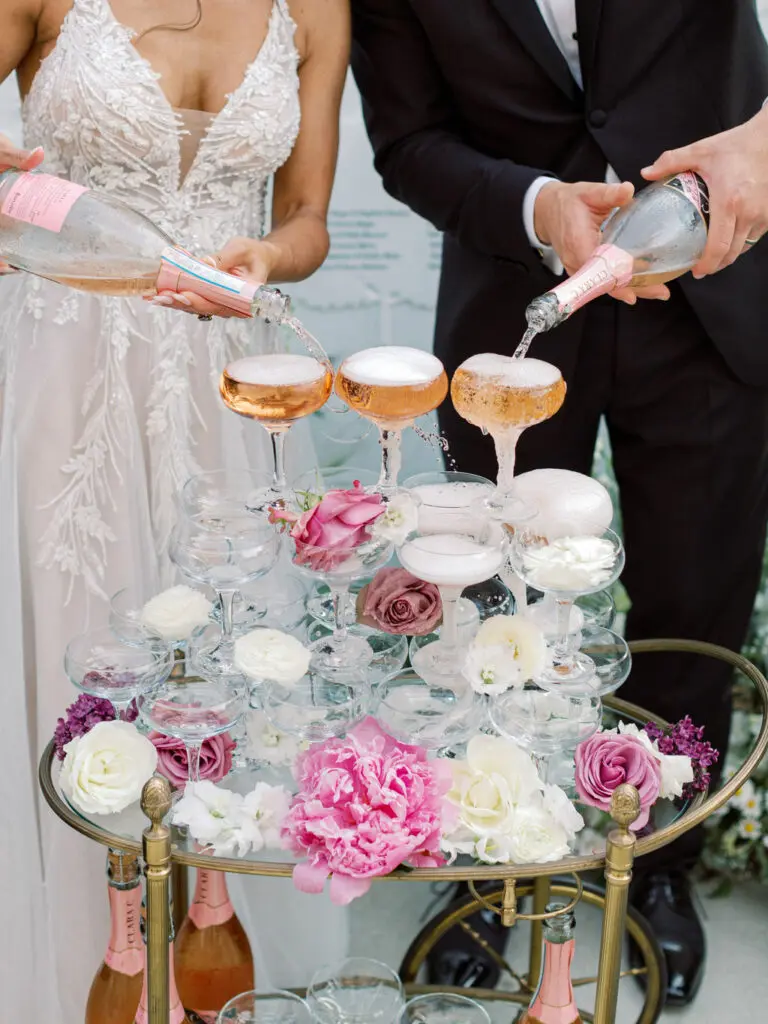 A bride and groom pour champagne into a tower of coupe glasses. The table's glass surface has some pink and white flowers.