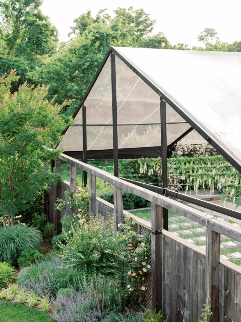 An outdoor greenhouse with a peaked roof and transparent panels stands surrounded by lush greenery. Various plants, shrubs, and flowers are visible in the well-maintained garden area around the greenhouse.
