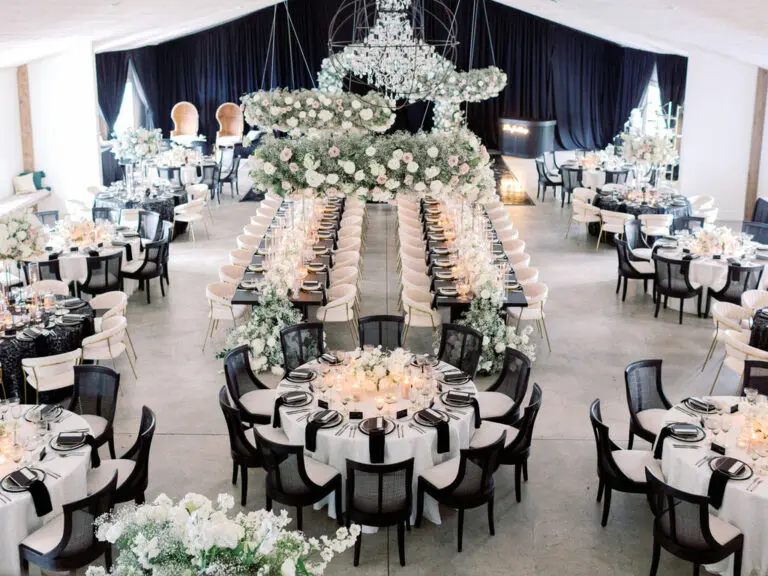 An elegant wedding reception setup features long rectangular tables adorned with white flowers and candles, surrounded by black chairs. In the center, a round table with similar decor is surrounded by black chairs. Floral arrangements hang overhead.