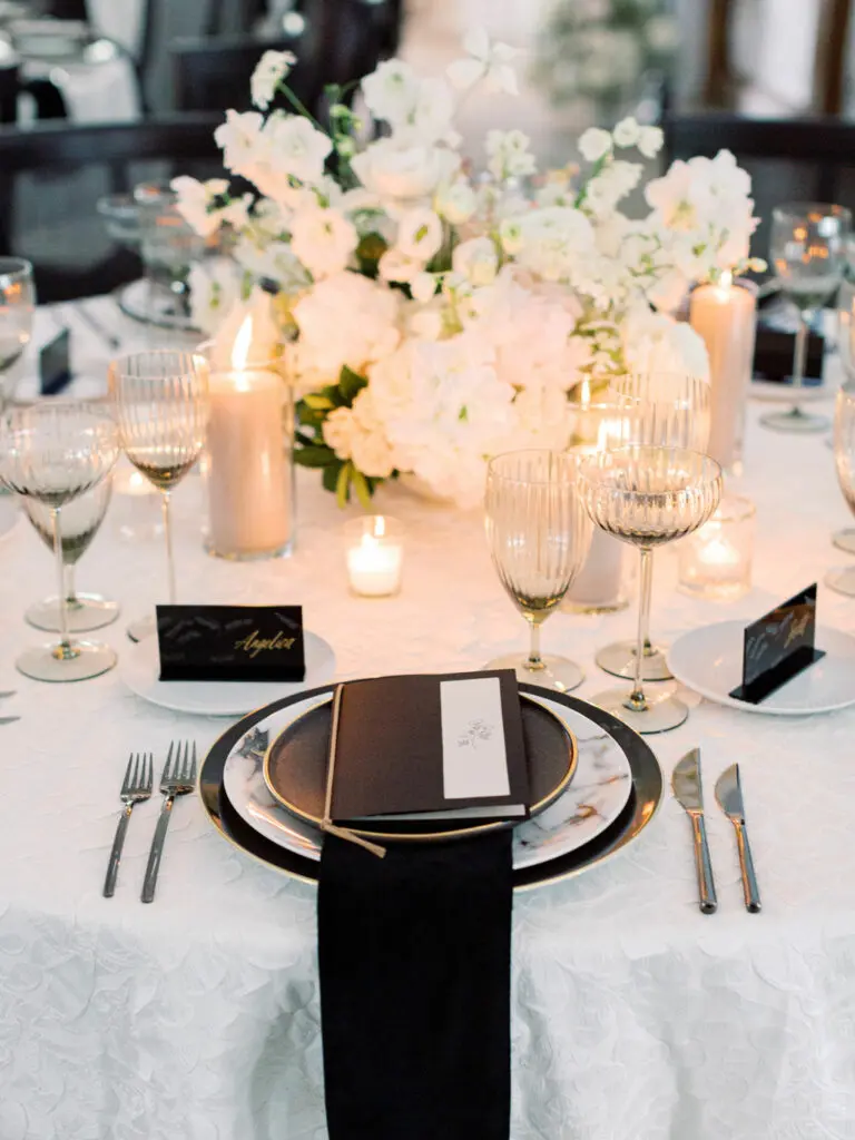 An elegant place setting features a short white floral centerpiece, votive candles, and a white tablecloth accented by black napkins.