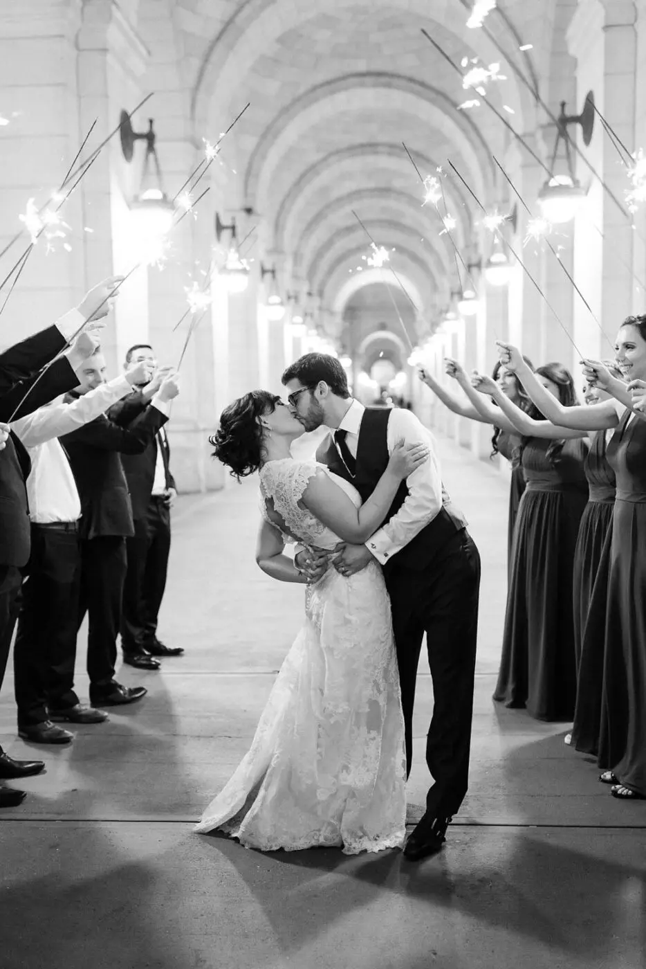A couple shares a kiss at their wedding, surrounded by friends and family holding sparklers under an archway. The bride wears a lace gown, and the groom is in a suit and tie. The scene is captured in black and white.