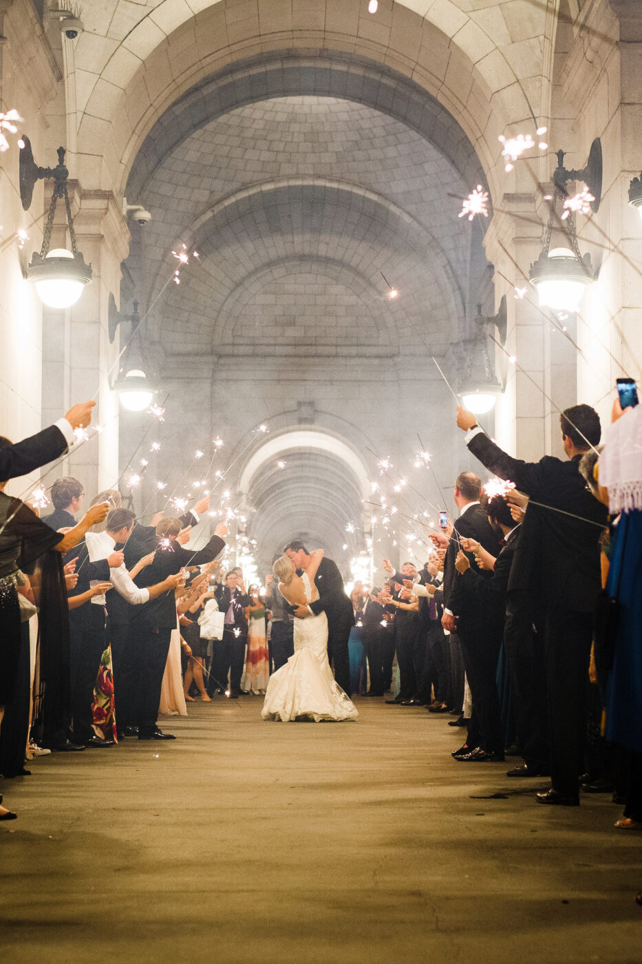 A bride and groom share a kiss under an outdoor archway at Union Station. Wedding guests hold sparklers on either side.