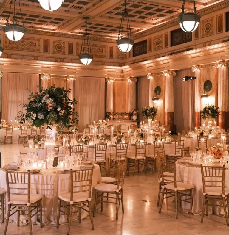 A grand ballroom set up for a wedding reception features ornately decorated ceilings and elegant chandeliers. Round tables, adorned with white tablecloths, floral arrangements, and candle centerpieces, are surrounded by gold chairs. A stage is visible in the background.