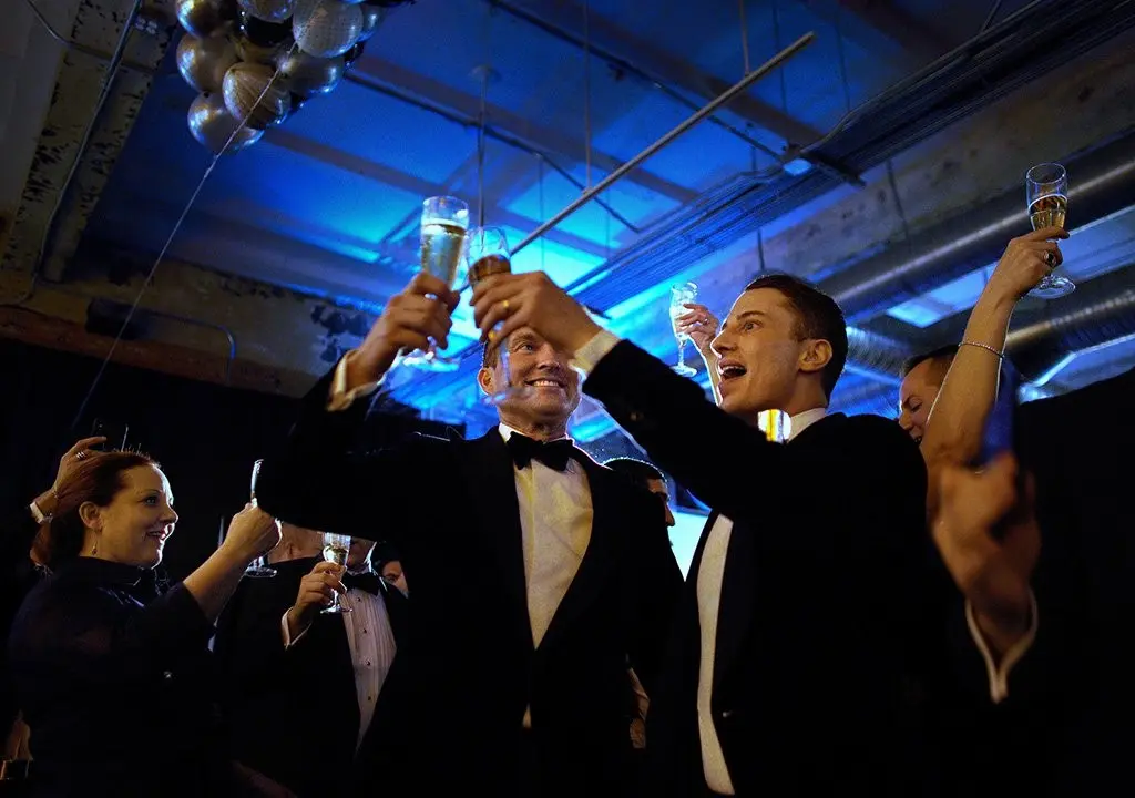 A group of people dressed in formal attire raise their champagne glasses for a toast in a festively lit room with blue lighting. Two men in the center, wearing tuxedos, smile and hold their glasses high, while other guests join in the celebration.