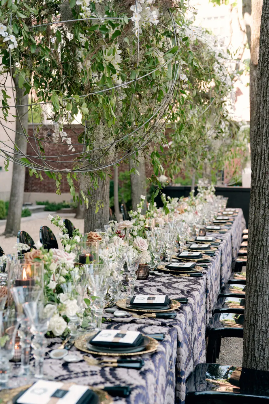 An long outdoor table is arranged with a navy and white patterned tablecloth. The table features floral arrangements of white and blush flowers, numerous wine glasses, and plates with navy napkins. Overhead, there are hanging floral decorations with greenery.
