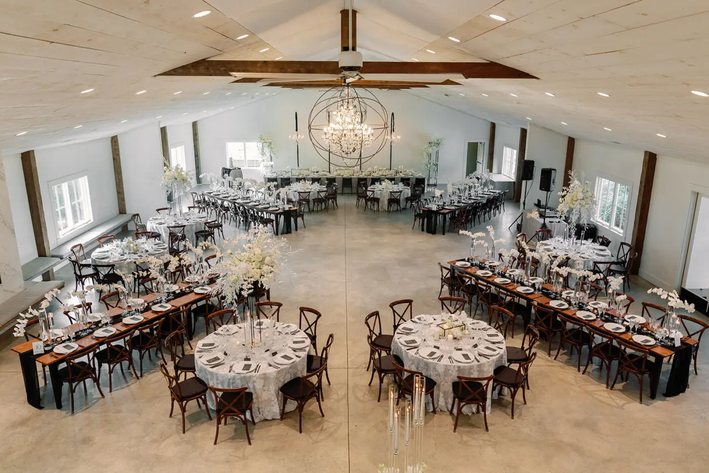 A spacious, elegant event hall with high ceilings and wooden beams is set for a formal dinner. Round and rectangular tables are adorned with white tablecloths, delicate floral centerpieces, and neatly arranged place settings. A large chandelier hangs in the center.
