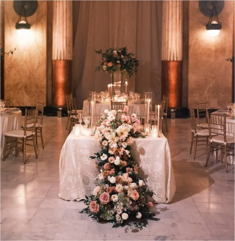 A table adorned with tall candles and a white tablecloth features light florals and greenery cascading toward the floor in an ornate banquet hall.