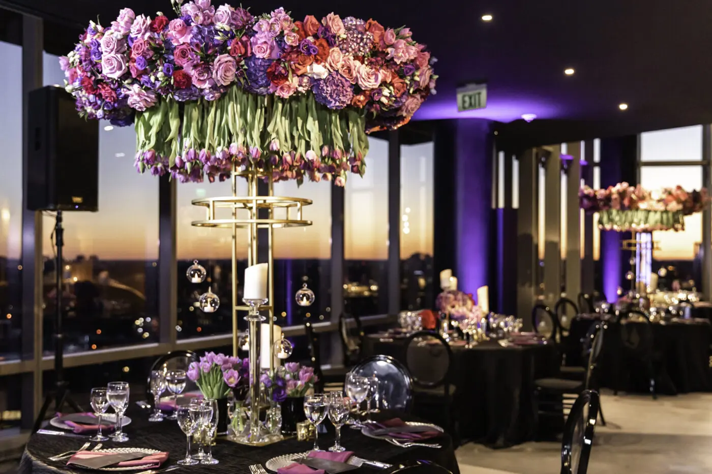 A decorated indoor event space is shown with tables set for dinner, featuring black chairs and black tablecloths. The tables feature large floral arrangements with purple and pink flowers. The sunset is visible through the large windows in the background that give an aerial view of the landscape.