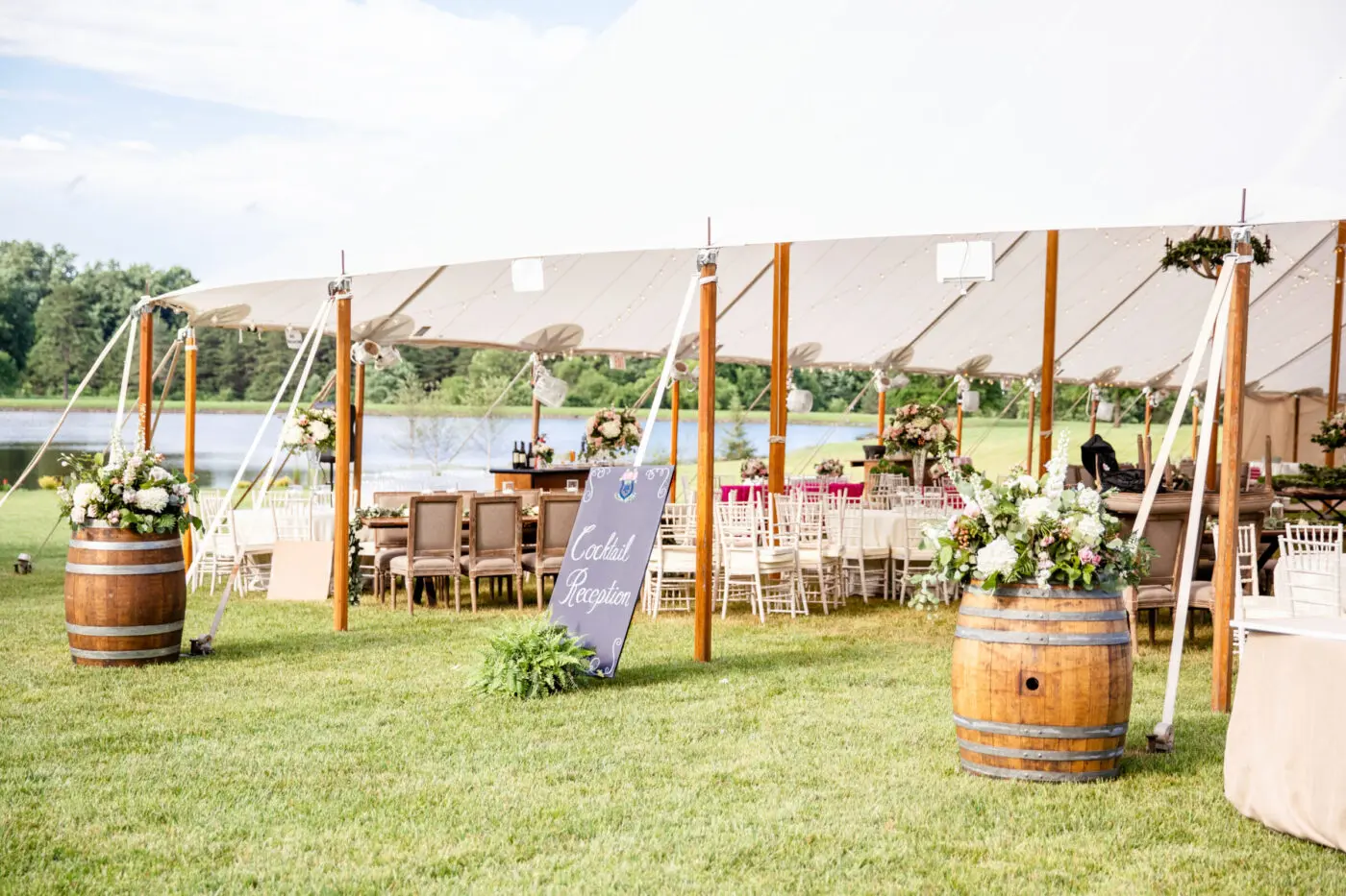 A white tent is set up on a grassy area near a lake for an outdoor event. Wooden barrels decorated with flowers stand at the entrance. Inside, there are elegantly set tables and chairs. A chalkboard sign reads
