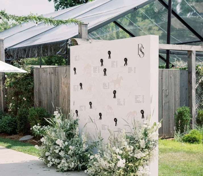 A large white seating chart board is adorned with black ribbon details and black lettering, placed outdoors next to lush floral arrangements with white and green flowers. The backdrop includes a wooden fence, greenery, and a structure with transparent ceiling panels.