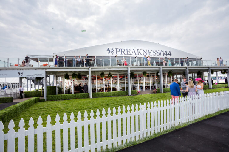 A wide-angle view of the Preakness event, showcasing a large, double-decked structure with the event logo. Attendees gather on the lawn and inside the structure, with a track visible to the right and a partly cloudy sky overhead.