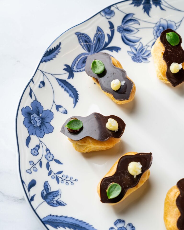 A decorative plate with a blue floral pattern holds six small pastry desserts. Each pastry is topped with shiny chocolate glaze, a dollop of cream, and a green leaf garnish. The plate is placed on a white marble surface.