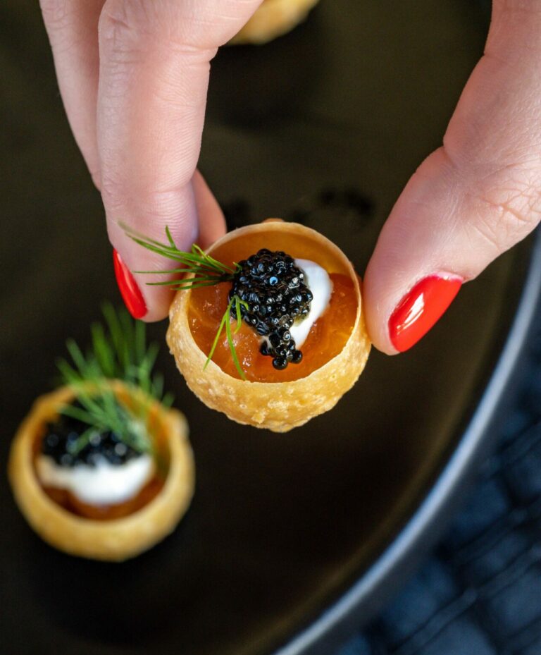 A hand with red nail polish holds a small, elegant canapé topped with sour cream, black caviar, and garnished with a dill sprig. More canapés rest on a black plate in the background.