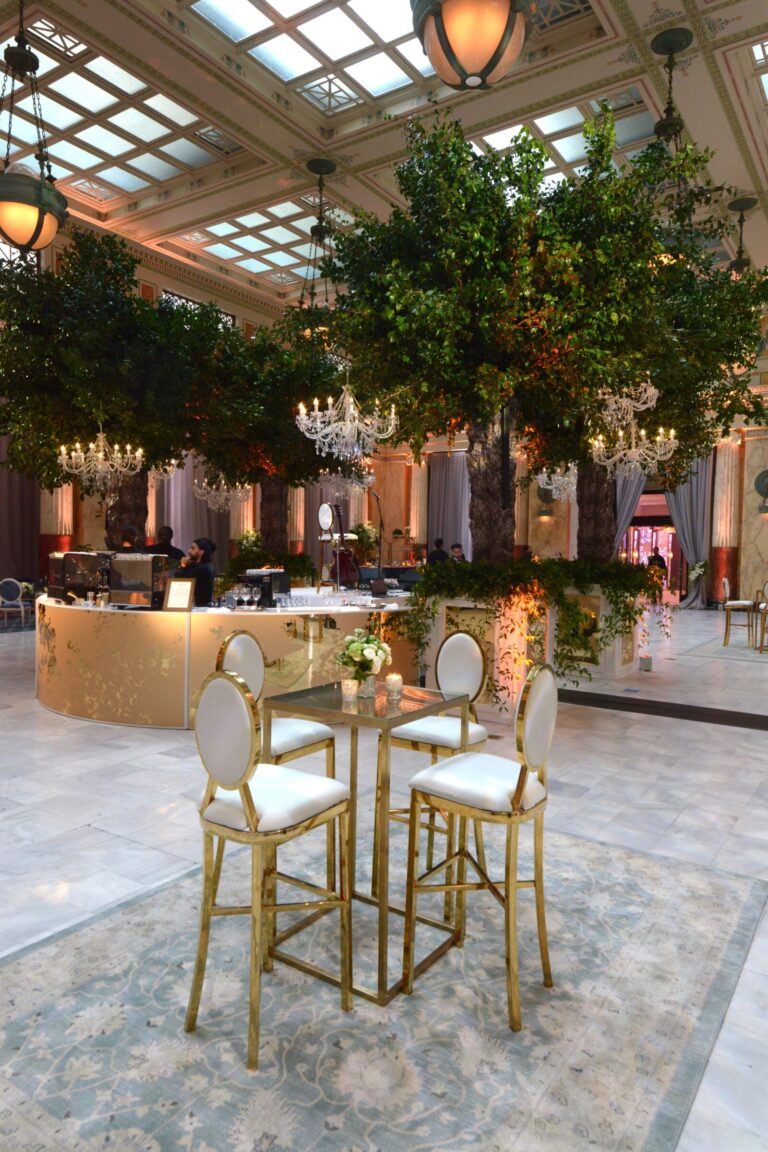 A small cocktail table with four chairs sits in the foreground of a brand event hall adorned with greenery and chandeliers suspended from the ceiling.