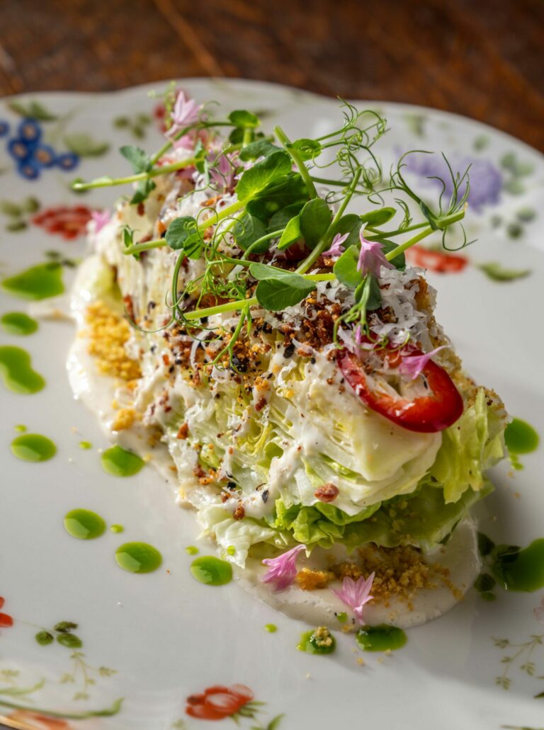 A beautifully plated gourmet salad featuring a wedge of lettuce topped with dressing, grated cheese, microgreens, and edible flowers arranged on a decorative floral plate. Bright green sauce dots surround the salad on the wooden table surface.