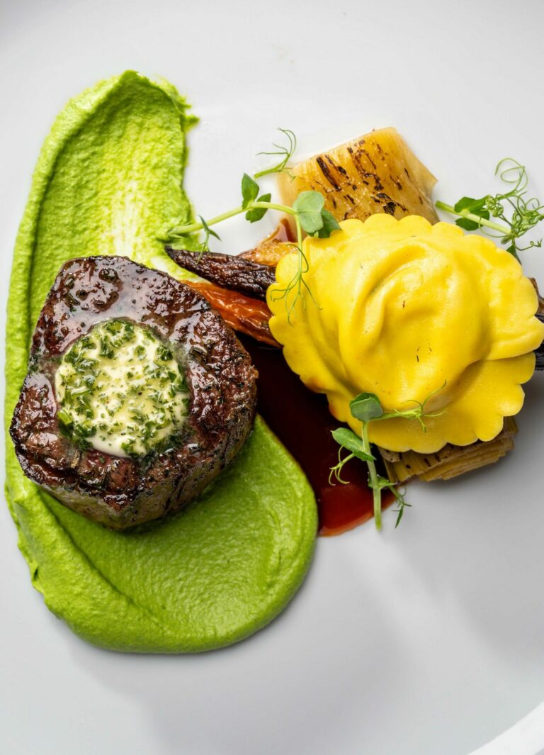 A gourmet dish featuring a seared piece of steak topped with herb butter, a small heap of grilled vegetables, a yellow ravioli with delicate garnish, all set on a smear of vibrant green purée on a white plate.