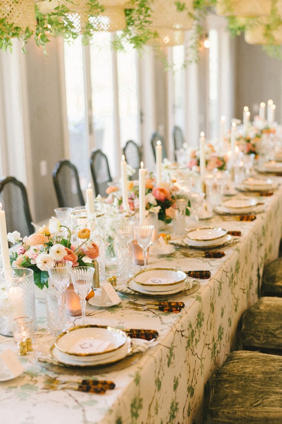 A long, elegantly set dining table with floral centerpieces, tall candles, and gold-rimmed plates. Crystal glasses and ornate cutlery are perfectly arranged, and green decorative foliage hangs above the table.