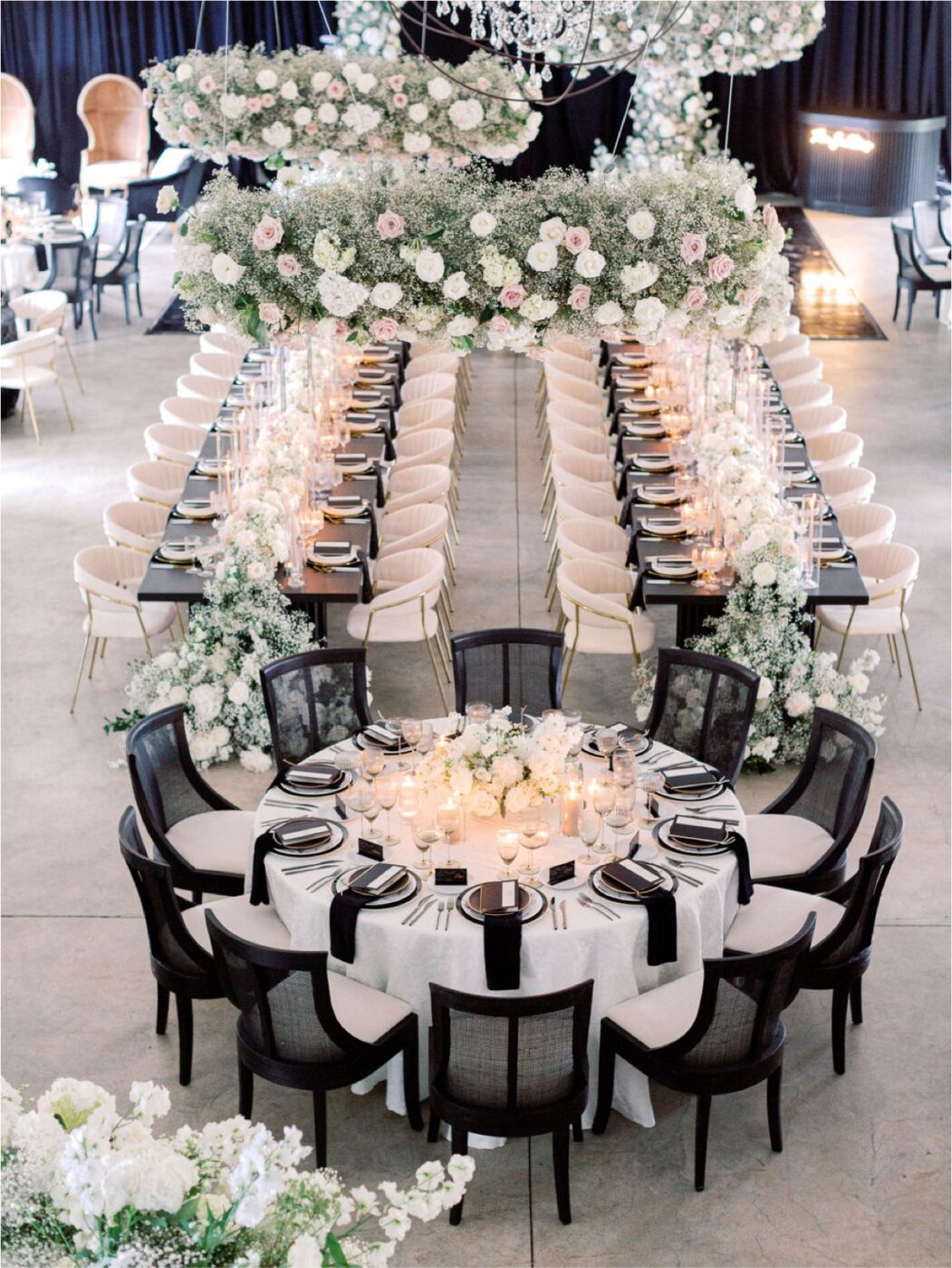 An elegant wedding reception setup features long rectangular tables adorned with white flowers and candles, surrounded by black chairs. In the center, a round table with similar decor is surrounded by black chairs. Floral arrangements hang overhead.