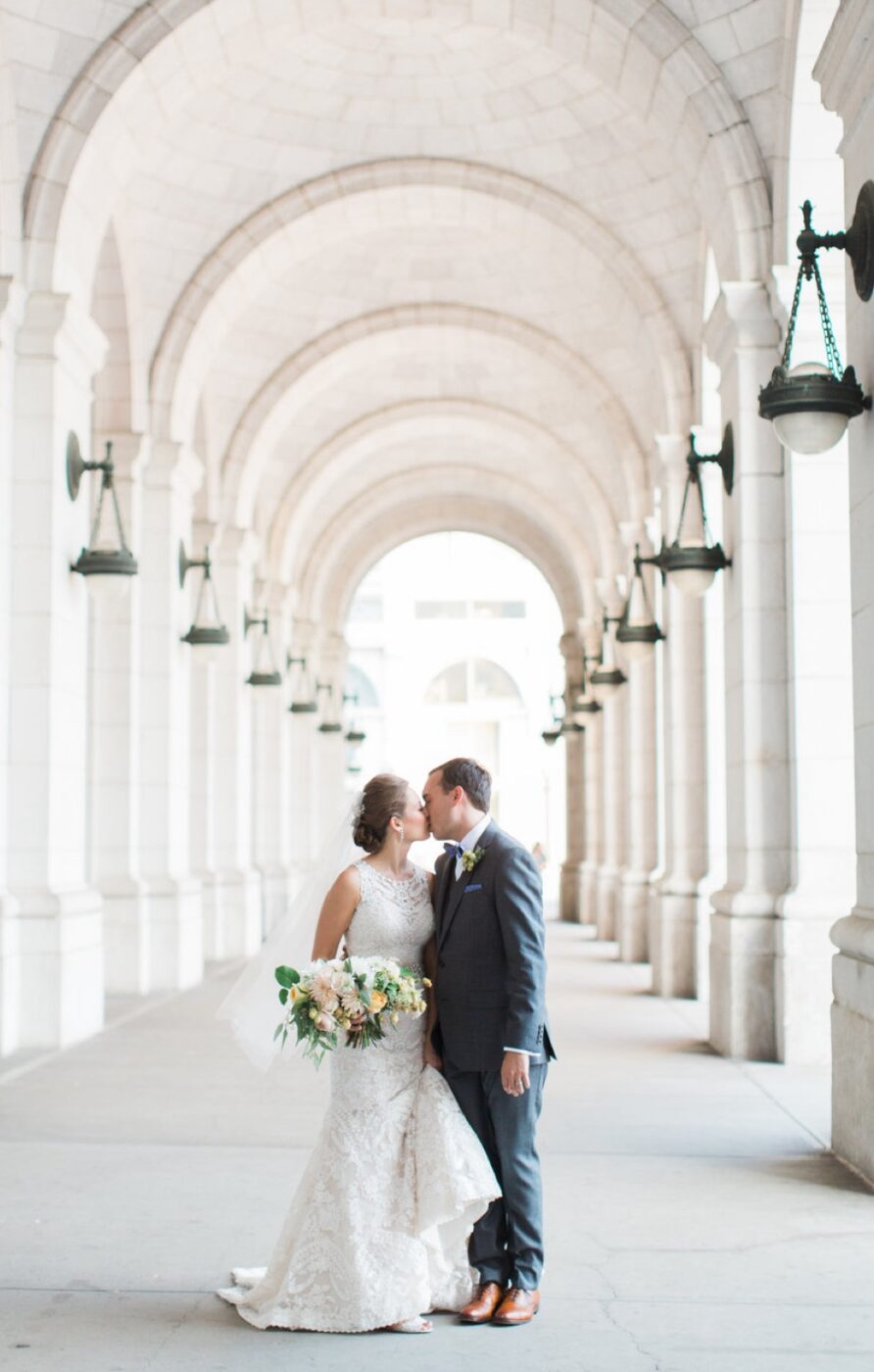 A couple shares a kiss under the beautifully arched hallway outside Union Station on their wedding day. The bride is in a white lace dress, holding a bouquet, while the groom is in a gray suit. Hanging lanterns and the repeating arches create a sense of depth and elegance in the background.